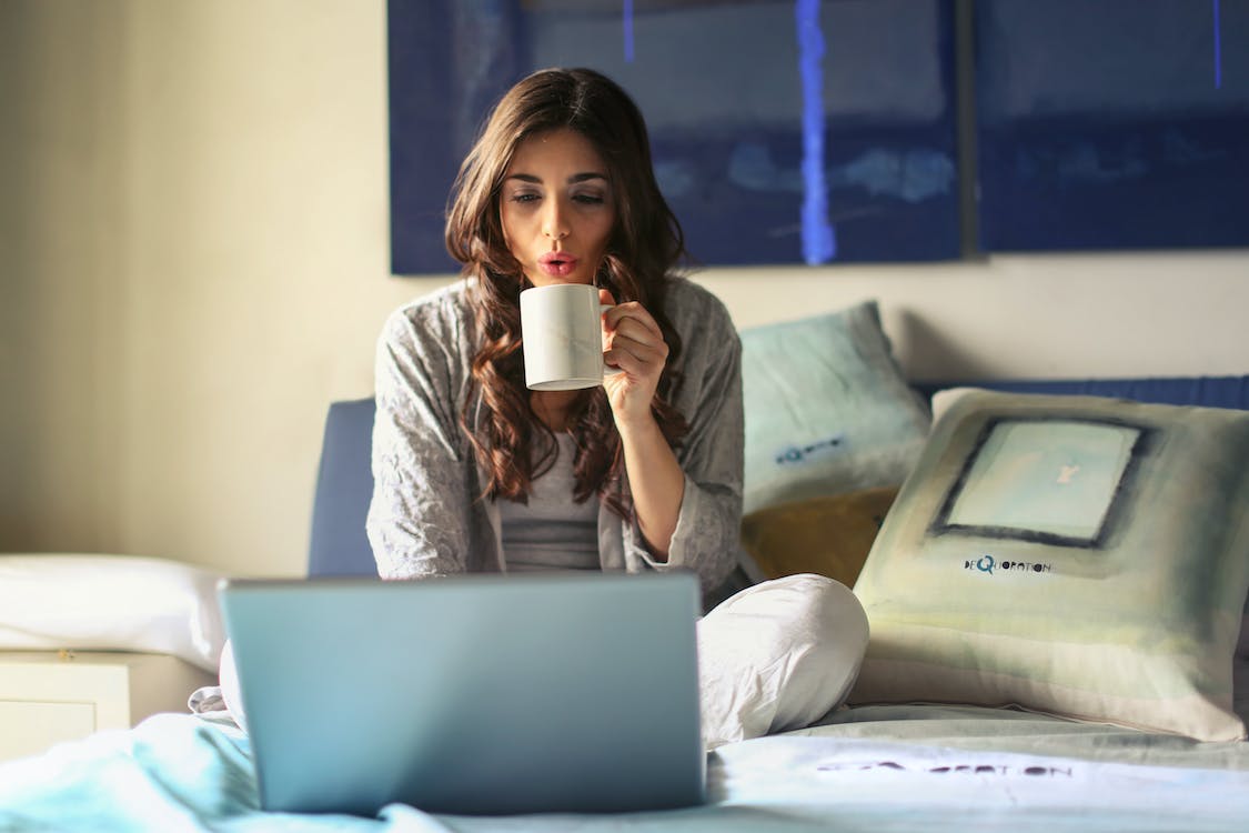 Free Woman in Grey Jacket Sits on Bed Uses Grey Laptop Stock Photo