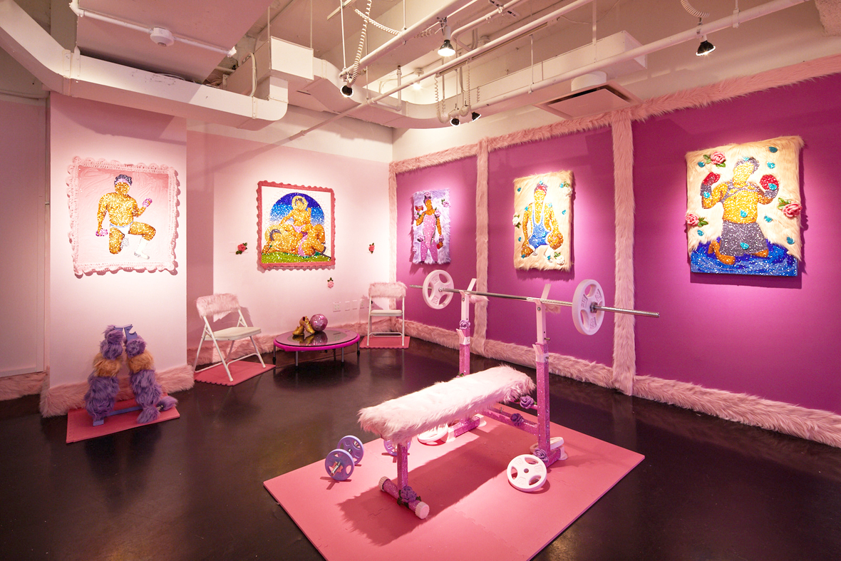 A well lit room with bright pink walls. On the walls are large images of semi-nude posed figures. In the room are pieces of excercise equipment, covered in pink fur. In the corner are two pink folding chairs and a low table. 