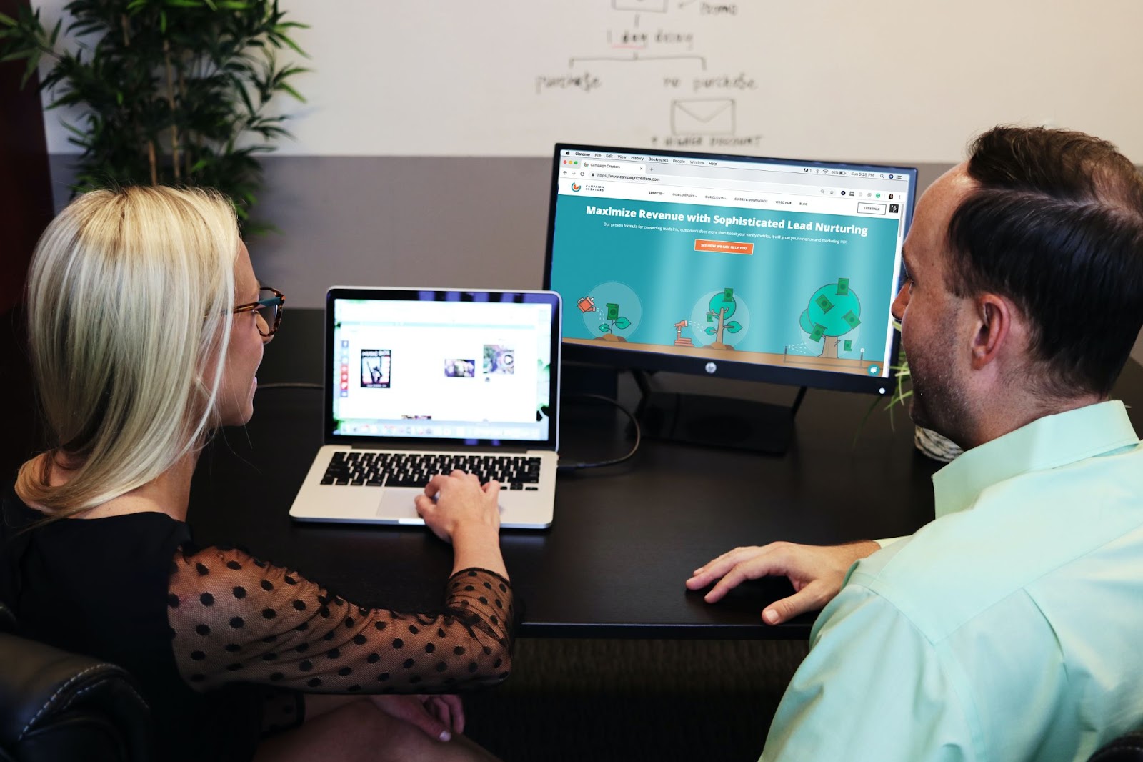 A man and woman sitting in front of a table with a laptop and computer monitor.