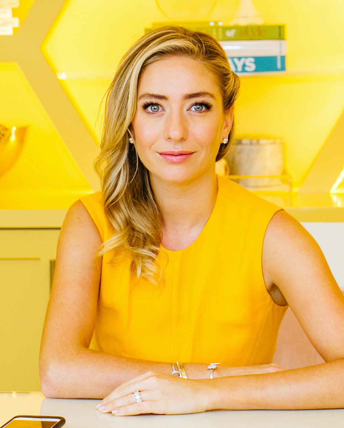 A portraiture photo of Whitney Wolfe Herd. She is in a yellow dress and is leaning both of her arms comfortably against the table in front of her. The background behind her is in the pattern of large honeycombs. 