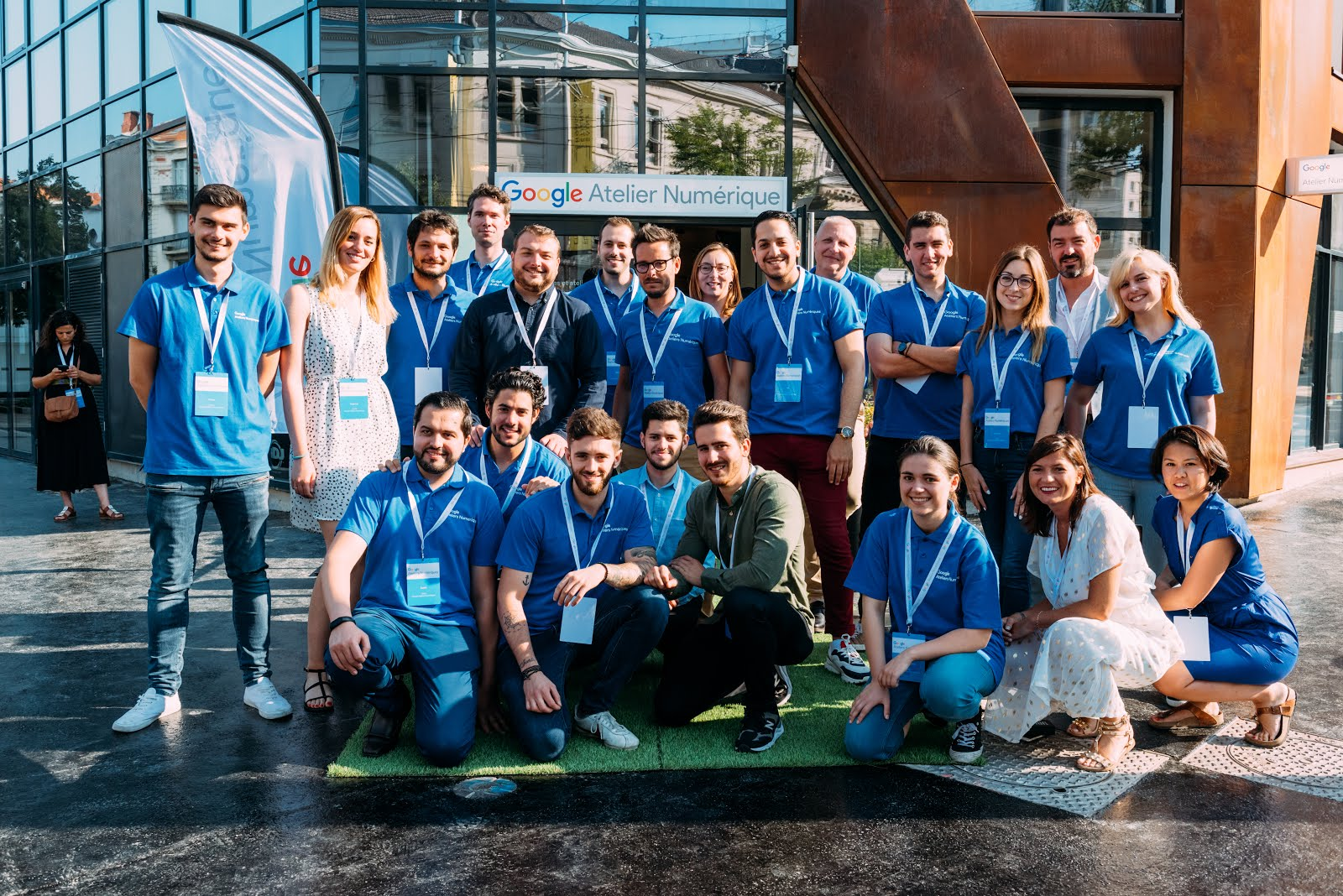 Un groupe d'environ 20 personnes, portant pour la plupart des chemises bleues et des badges assortis, souriant devant le bâtiment Google Atelier Numérique