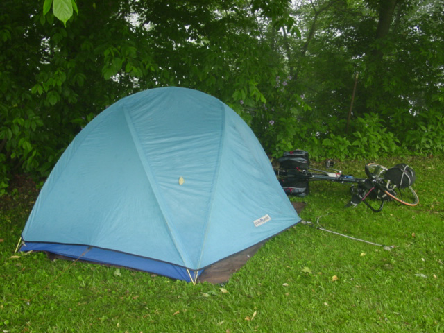 Blue rainfly over a tent against some trees with bicycle lying in the grass behind. 