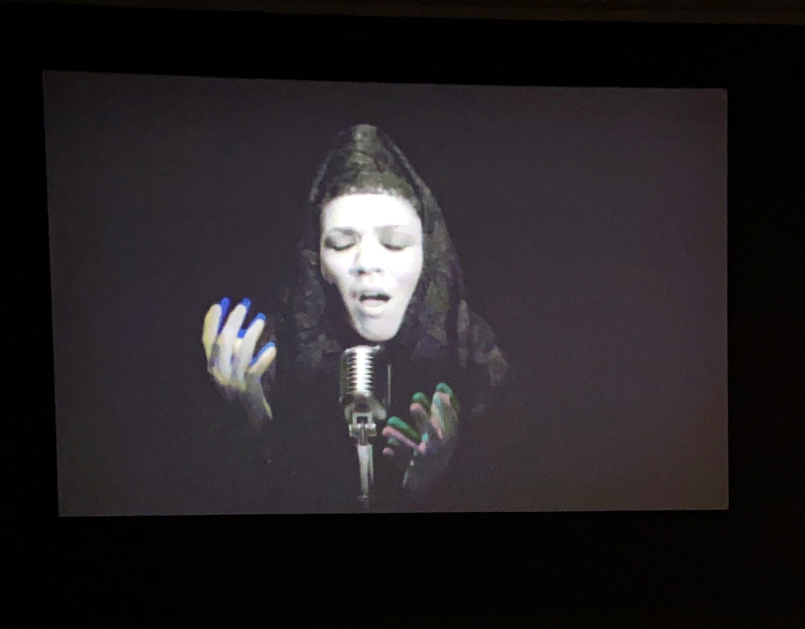 Image: Shirin Neshat, Turbulent (still), 1998, two-channel video installation, 16mm black and white film transferred to video. A photograph of a video projection on a black wall. Sussan Deyhim, an Iranian singer, is pictured from the chest up in front of a microphone. She wears all black, including a black veil over her head. Her face is exposed; her eyes are closed and her mouth is open in song. Her hands are lifted toward the microphone. There is slight discoloration and echo of her hands as they were photographed while in motion. Photo by Jessica Hammie. 