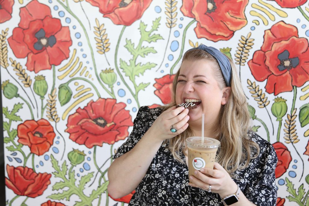 Erin Peterson enjoys a pastry and iced coffee in front of a colorful flower wall at Made With Love Bakery.