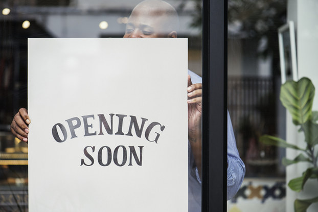 This is a young black male inside his business office hanging a banner that reads 'opening soon'. 