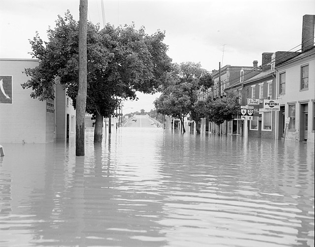 A black and white photo shows standing water on a commercial street with shops and buildings.