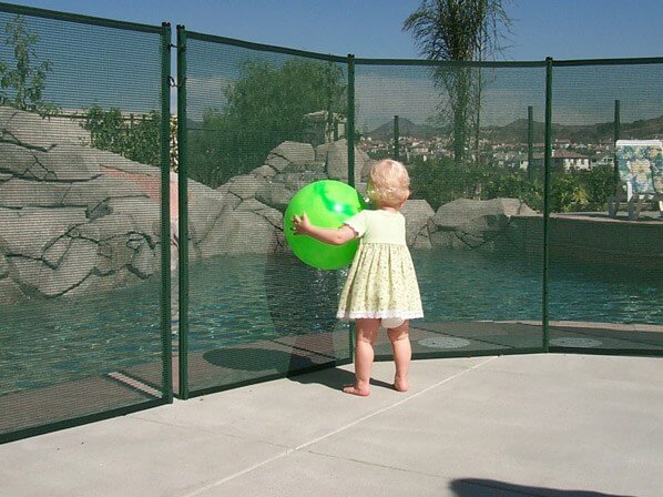 toddler standing against a pool fence holding a green ball