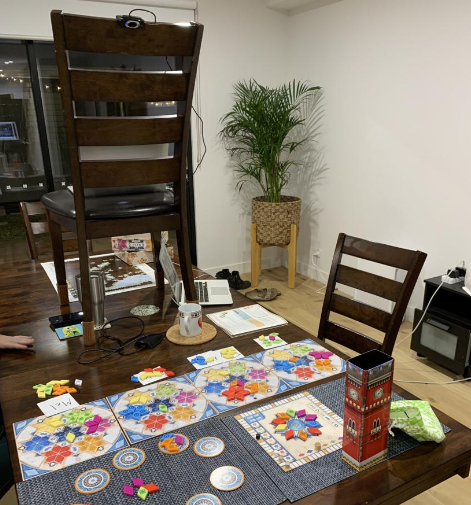 Doug's webcam setup for broadcasting and piloting the boardgame Azul over videochat. (Pictured here is the third game in the series, Azul Pavilion, not the original Azul, which we prefer). There are 5 boards on a table with counters, pieces of paper with player's names on them, and a chair ON the table which has a laptop on it with camera.