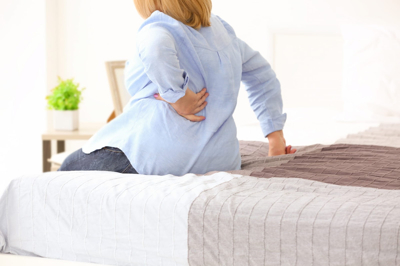  A woman in a sweater sits on a bed while holding back due to pain when taking a deep breath.