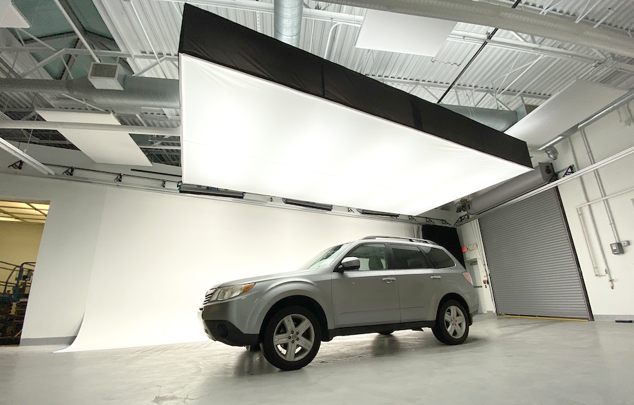 A sound stage big enough to fit a Subaru mini-SUV inside. A massive box light is suspended above the car with a white cyclorama in the background. There is a large roller-style door that the car could use to get into the studio.