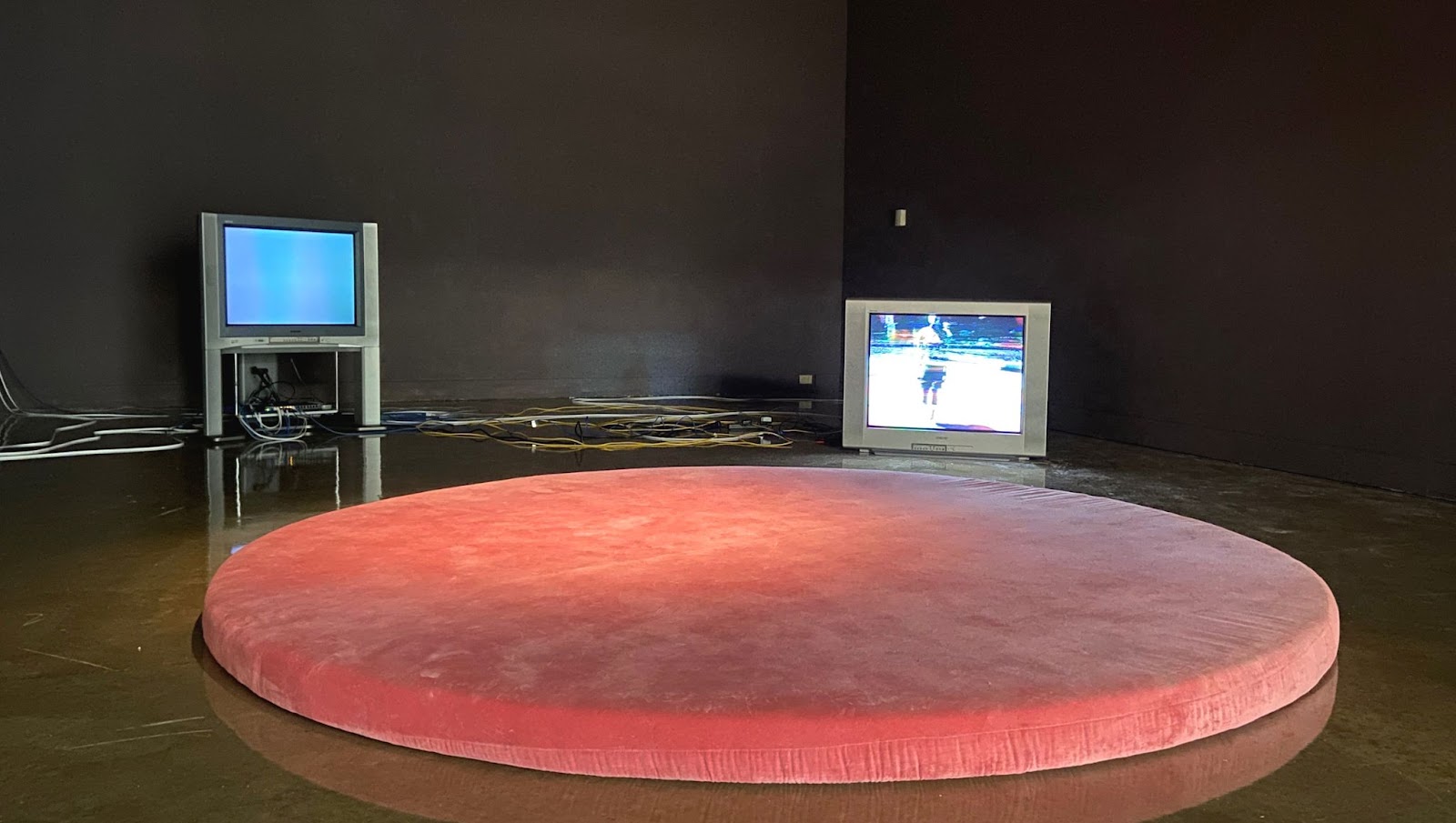 Image: Pipilotti Rist, (Enlastungen) Pipilottis Fehler (still), 1988, single channel video. Installation at the Tarble Arts Center, 2021. A large, circular, pink, upholstered platform sits on a concrete floor. Behind the platform are two 1990s-era televisions: on the right, the television sits on the floor. On the screen is a disrupted image of a person. On the left, a television is raised on a platform. The screen is blue. Several wires are on the floor behind the televisions. Photo by Jessica Hammie.