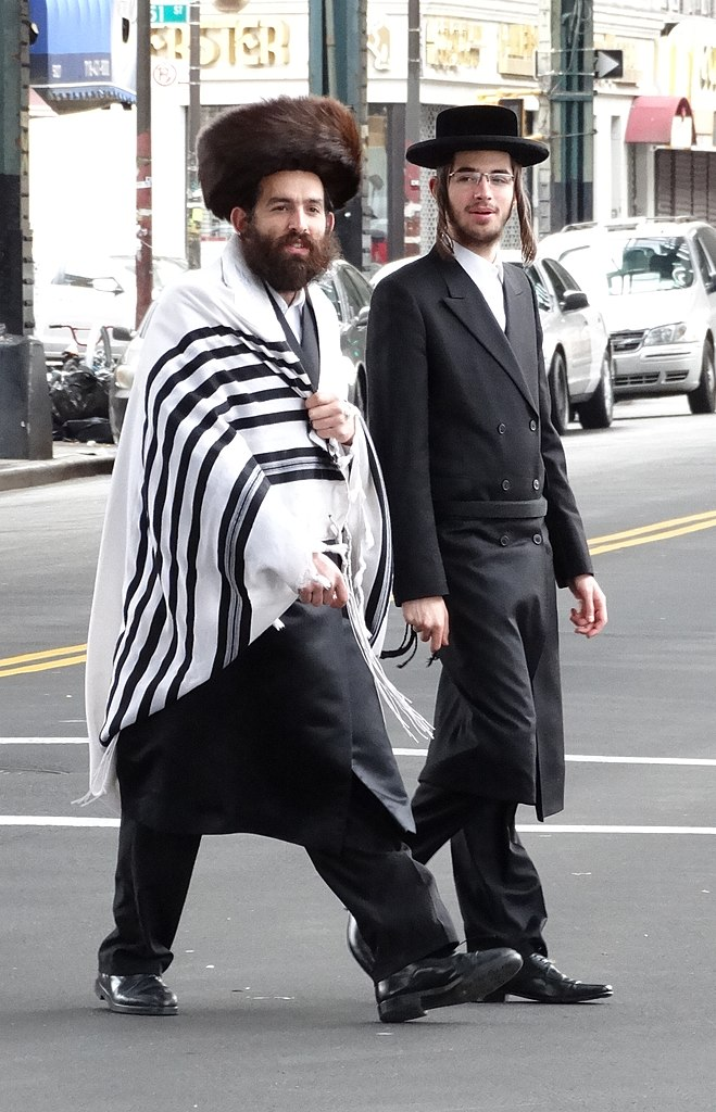 Two religious Jewish men walking down the street. One is wearing a tallit and a streimel.
