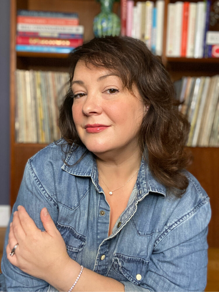 Alison Gary of Wardrobe Oxygen wearing a denim shirt sitting in front of a bookcase holding record albums. She is wearing James Allen diamond stud earrings, tennis bracelet, and pendant necklace