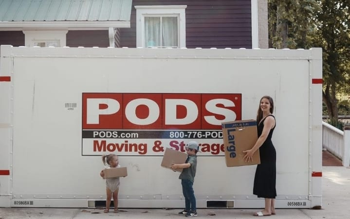 Katherine Tuttle and her two children are standing in front of their PODS container, each holding a box.