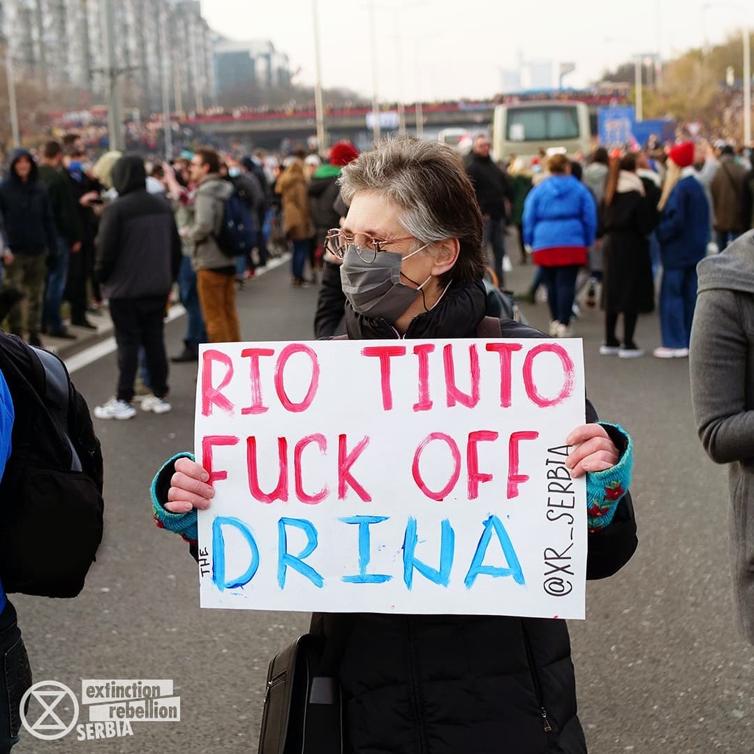 An elderly woman holds a sign telling rio tinto to fuck off. She stands with others in a main road in Belgrade.