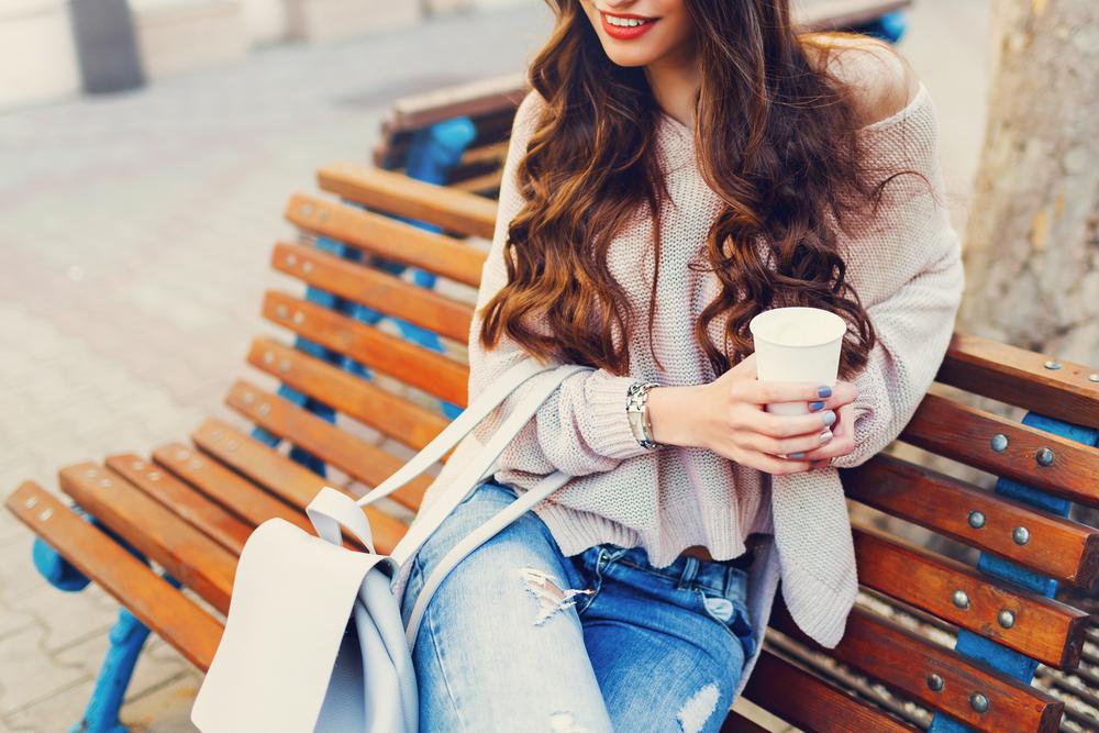  Outdoor fashion details. Sunny lifestyle  image of   stylish young  attractive woman in cozy knitted sweater  , holding cup of  hot  beverage , sitting on wooden  city bench.