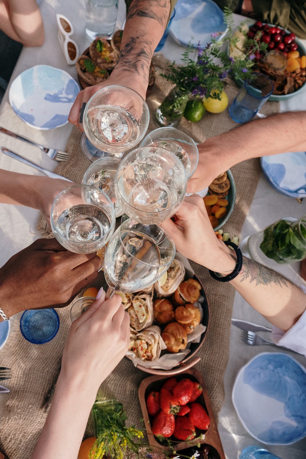 A group of six hands clinking glasses of water over a table of mixed food items. 