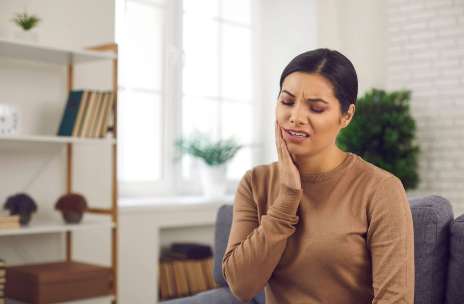 A lady in a brown-coloured long-sleeve shirt is sitting on a sofa with her right-hand touching her left face which is swelling because of smoking.