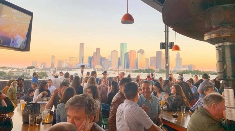 Dozens of people enjoying Houston nightlife at a rooftop bar as the sun sets behind the city skyline.