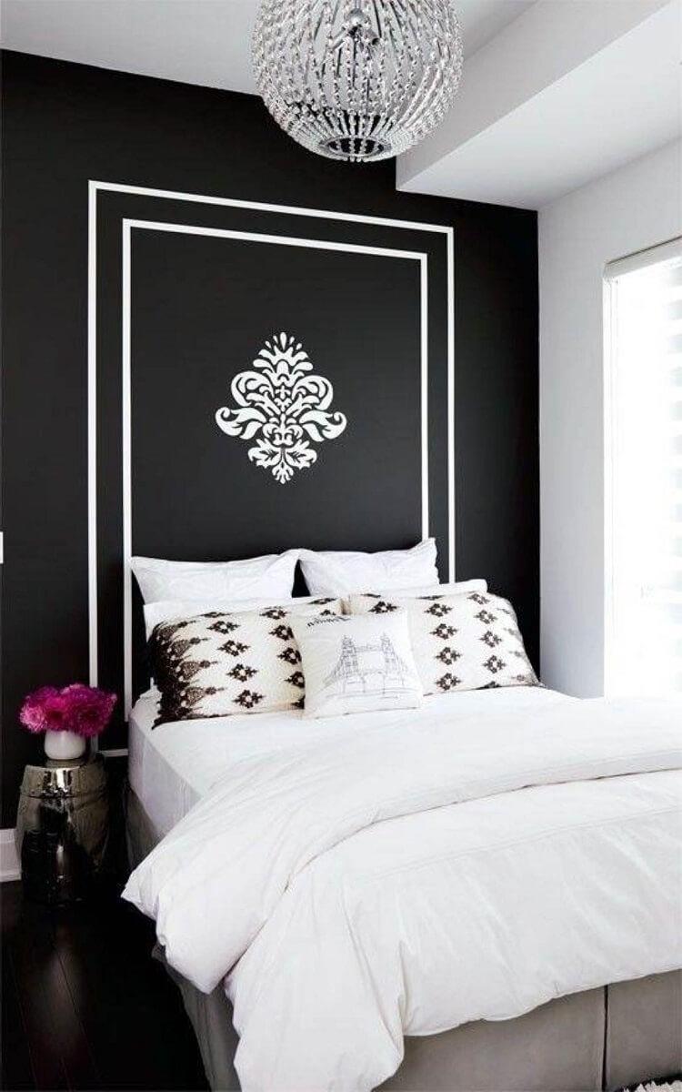 Quarto com cama de casal com parede da cabeceira preta com brasão branco no centro,  mesinha de canto preta com vaso de flor e piso de madeira.