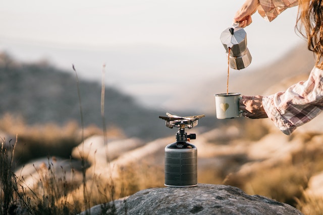 A woman pouring coffee from a coffee pot. Besides, the is a grey stove placed on a stone. These are essentials of a complete kayak camping gear