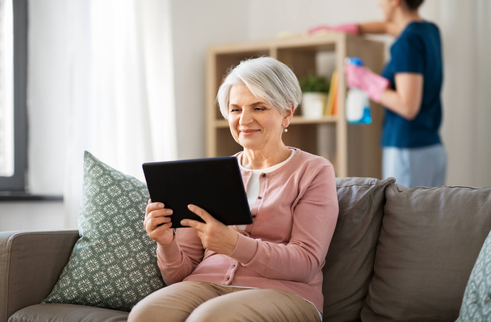 A senior woman sits on a couch while looking at a tablet while in the background someone dusts a cabinet.