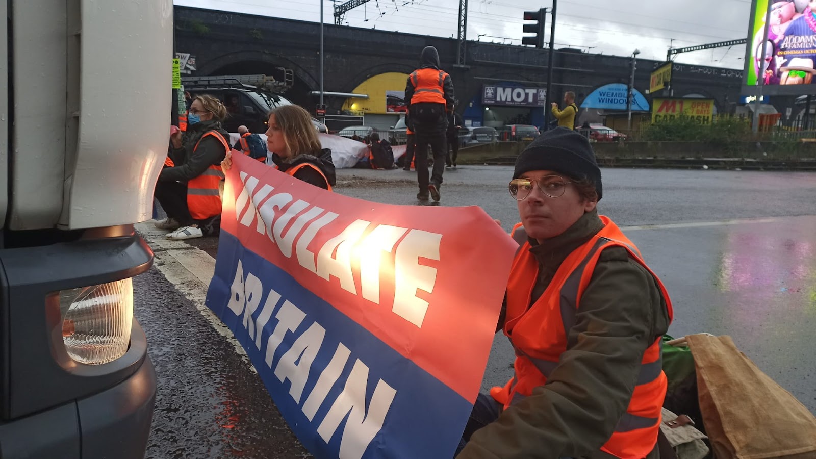 A young protester kneels on a road holding an insulate britain banner. A huge whie lorry is parked right in front of him. He looks to camera.