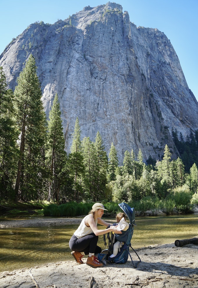 A person is bending down to help a toddler in a child carrier next to a river at the base of a tall rock mountain.