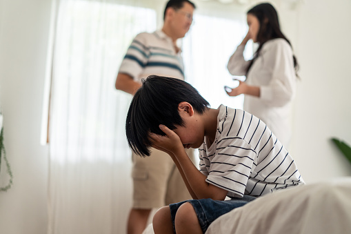 Two parents standing and having an argument while their son is sitting on the side of the bed and having his head covered with his hand looking sad. You can also get help for couples counseling in Los Angeles, CA and with online therapy in California.  91344