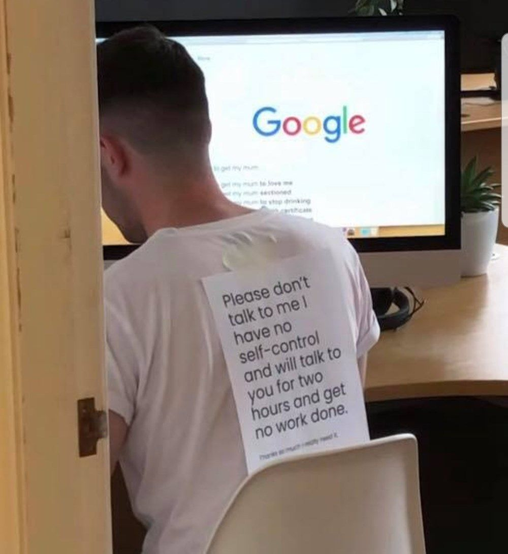 Man at a desk with a piece of paper taped to his back saying, “Please don’t talk to me I have no self-control and will talk to you for two hours and get no work done.”