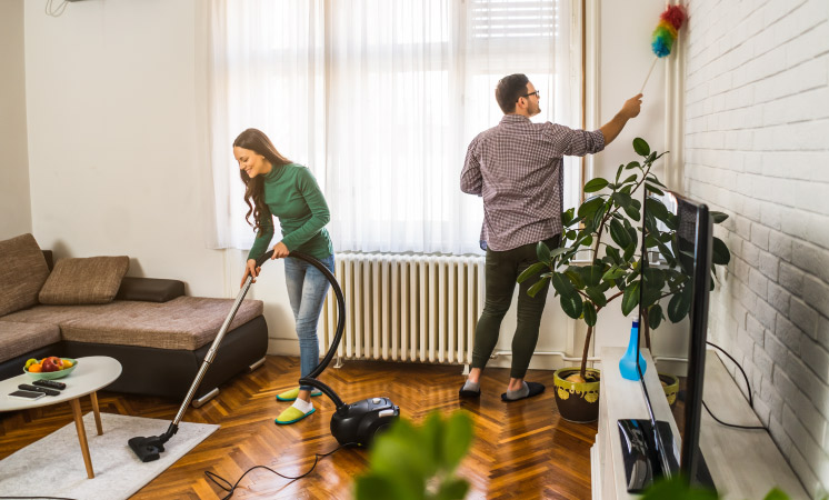 A couple cleaning their living room, she vacuums an area rug and he dusts a wall corner