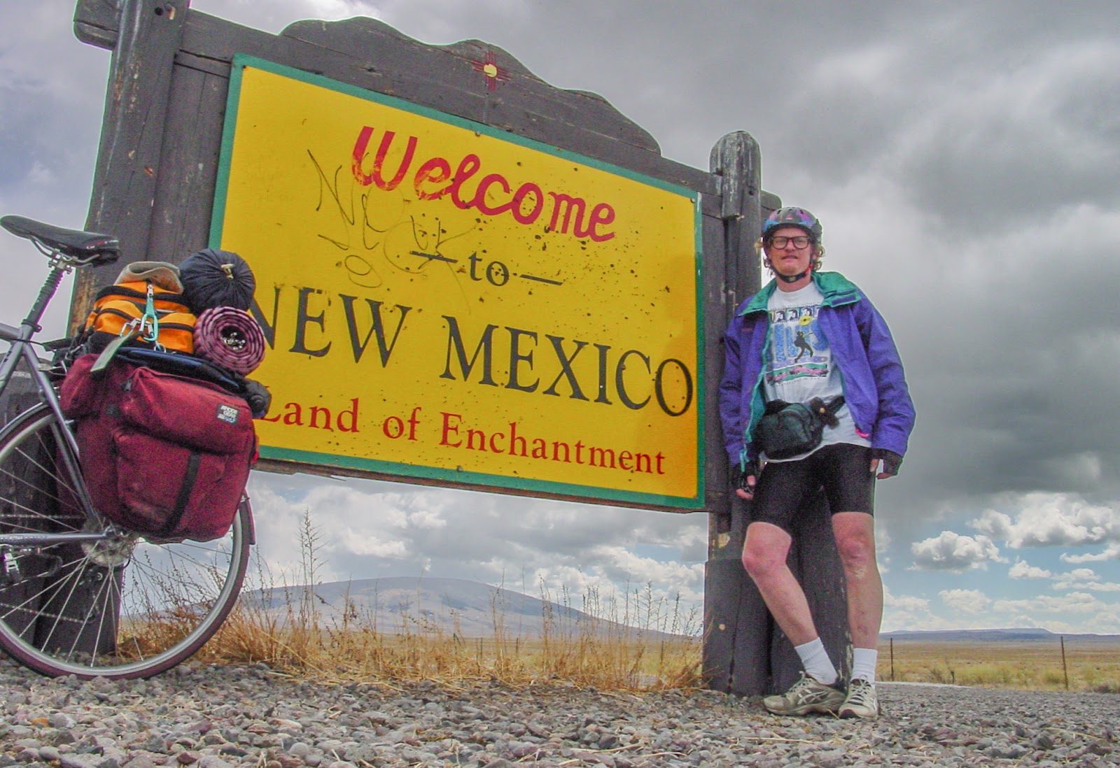 Cyclist leaning on a sign that says: Welcome to NEW MEXICO Land of Enchantment. 