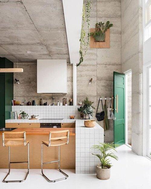 Cozinha com parede de cimento queimado, porta verde, piso porcelanato branco, bancada revestida de azulejo branco, bancada de madeira com bancos. 