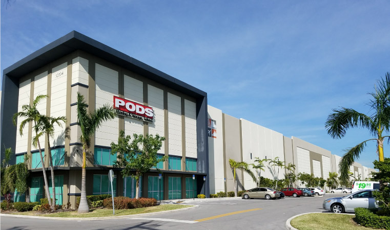 A PODS Storage Center in sunny Miami, Florida. Palm trees stand outside the facility's aqua doors and cars bake in the sun.
