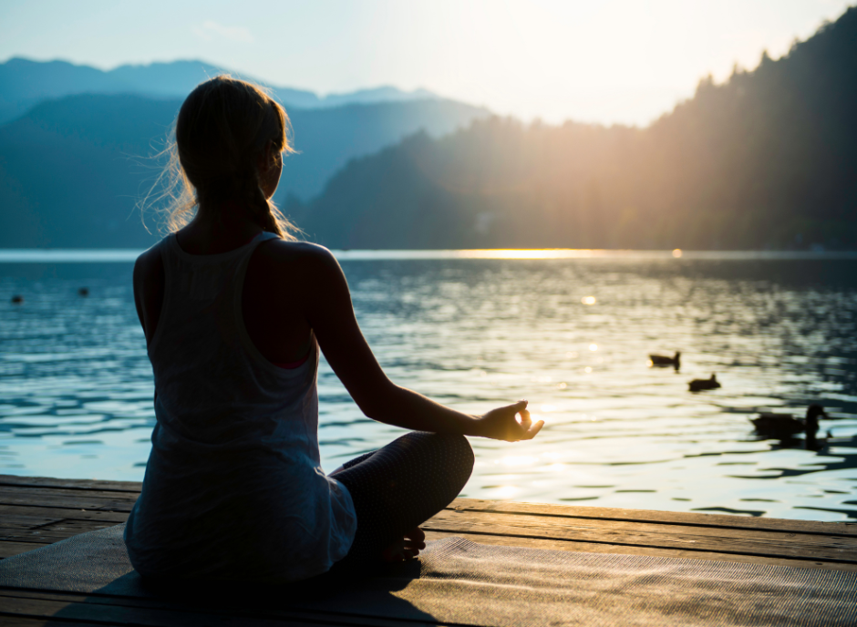 Woman sitting in mediation, overlooking a tranquil lake and mountains.