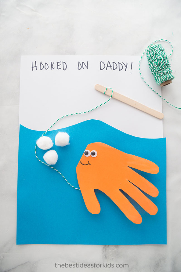 A fish handprint card with an orange fish attached to a fishing rod made with a piece of green and white string. There is a spool of green and white string beside the card.