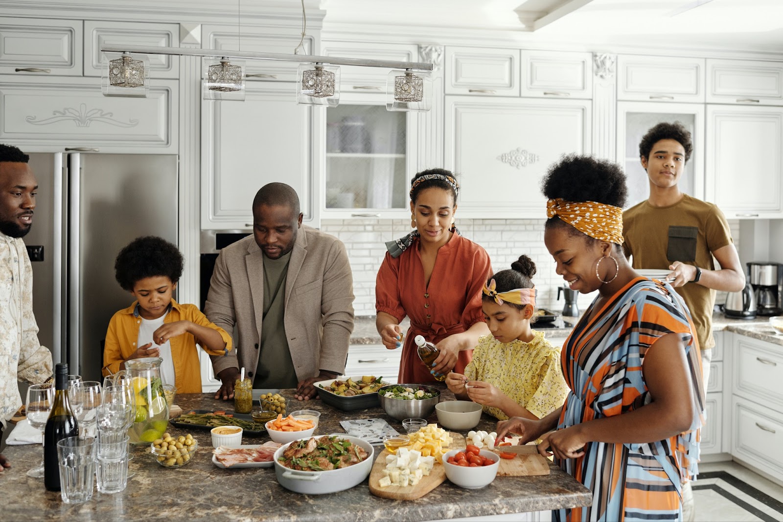 A family, including two adult men, one adult woman, two teenagers, and two younger children, gather around a kitchen island to prepare food.