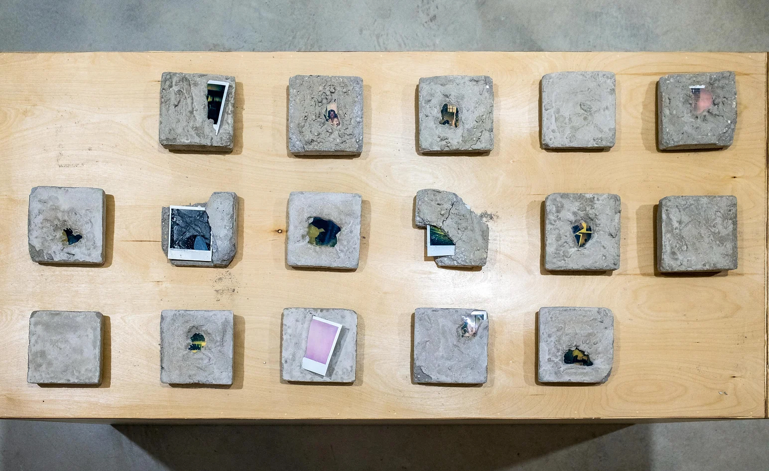 Image: An installation view of the 2002 work, In Remembrance of Us. Fifteen squares of concrete are seen from above and arranged in three rows on a wooden table. Each square is different and many have Polaroid photographs pressed into them. Photo courtesy of Sibyl Gallery.