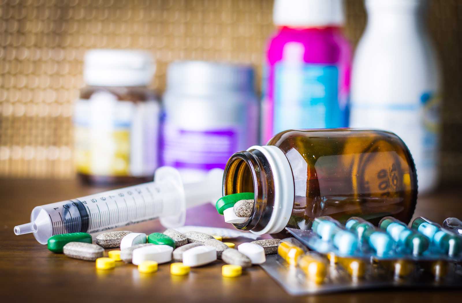 assortment of medication spilling onto wood table with bright pink and white bottles out of focus in the background