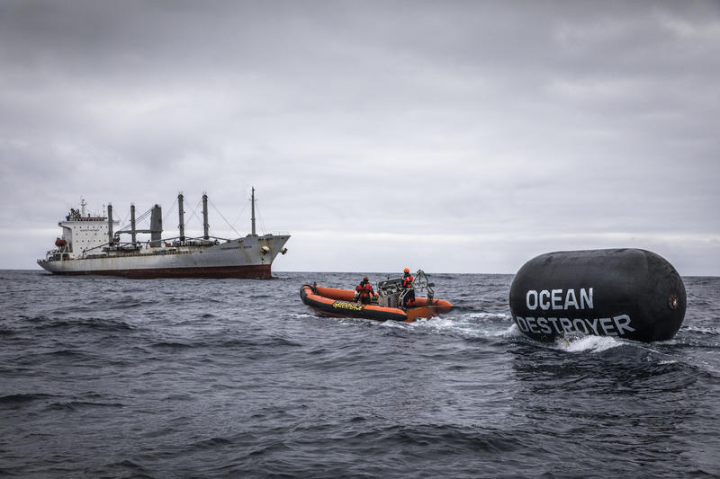 An orange dinghy heads towards a fishing boat, towing a big buoy with the words OCEAN DESTROYER painted on it