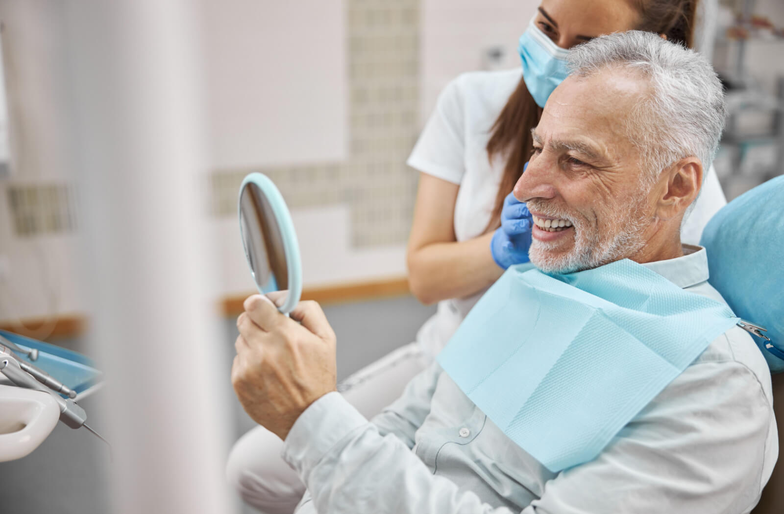 A senior man sitting in a dentist's chair, holding up a mirror and smiling with his dentist in the background