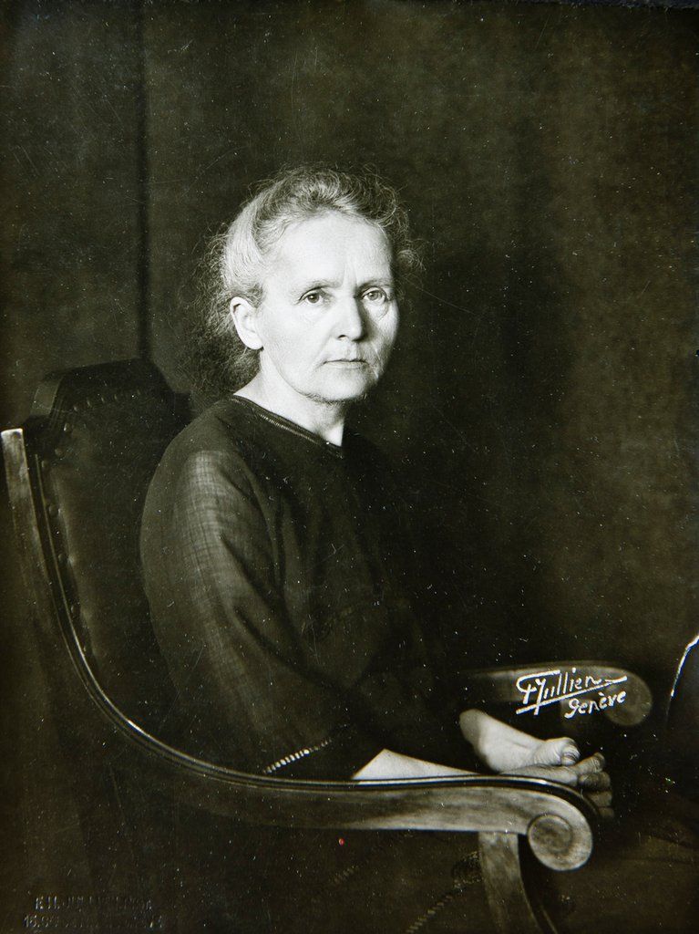Cientista Marie Curie, mulher branca e de cabelos presos, sentada em uma cadeira de madeira.