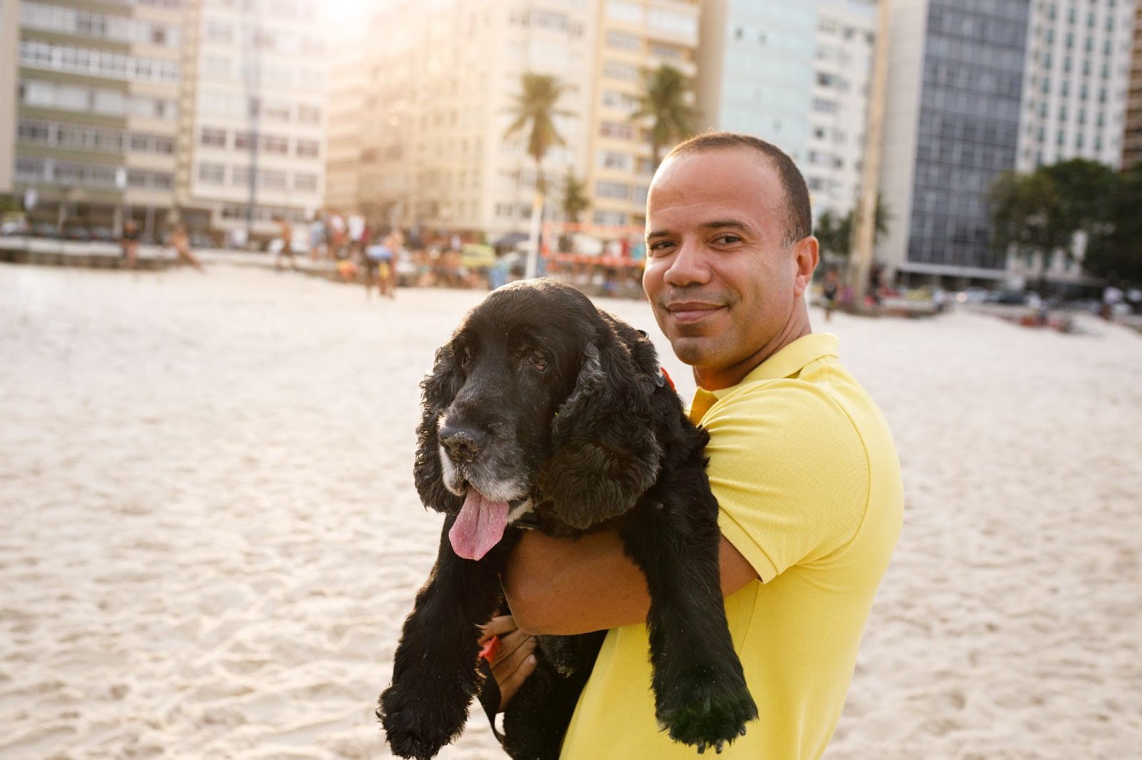 Na praia, homem sorrindo enquanto carrega cachorro preto no colo 