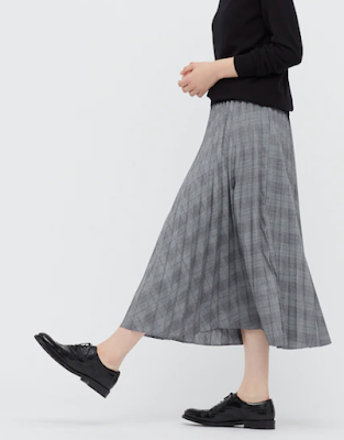Person wearing a gray long plaid accordion pleated skirt and black Oxford shoes, from the side, with one foot slightly raised in front of them.