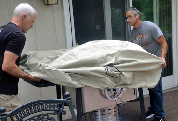 Two men put a tan cover over a shiny gas grill