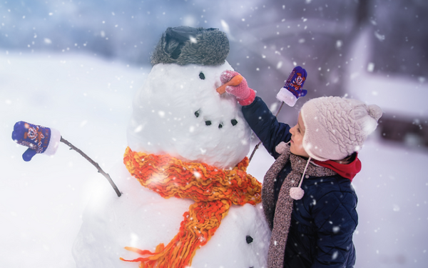 Young child outdoors in the snow wearing a jacket, scarf, and hat placing a carrot in a snowman’s face