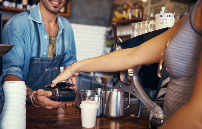 A woman using her phone to pay for a coffee. The barista is smiling and holding out the payment terminal. 