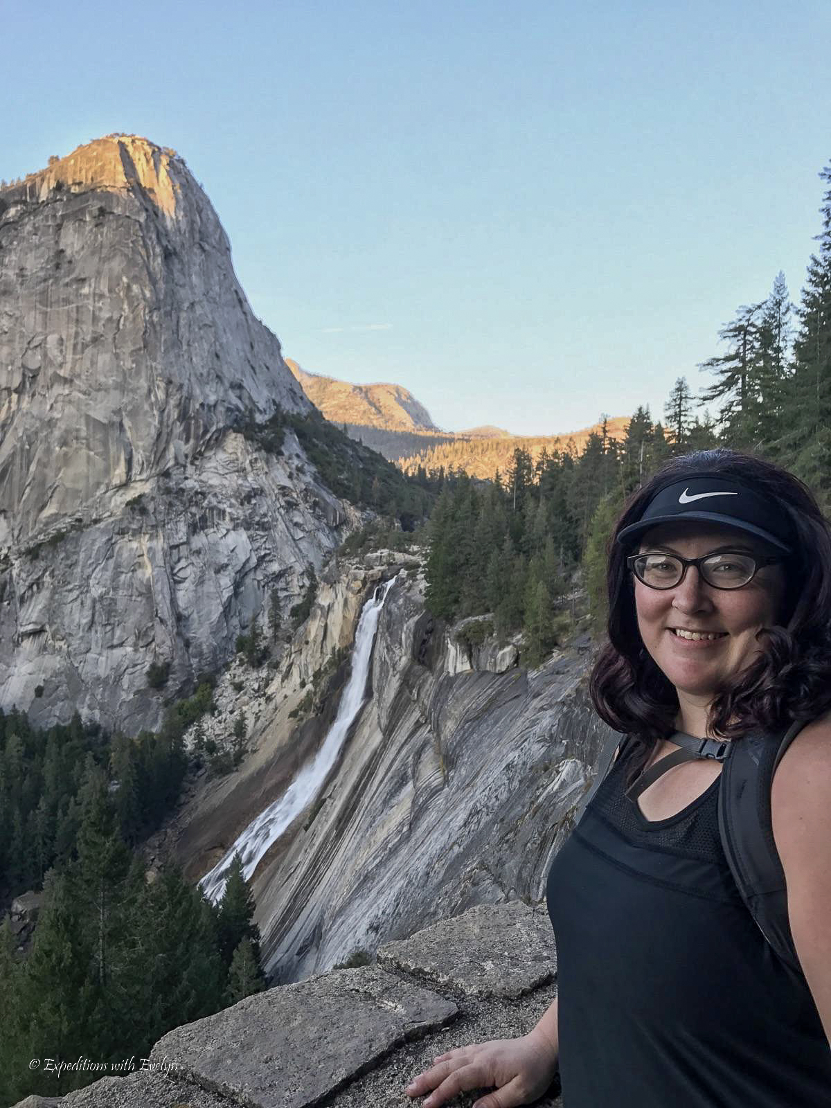 A female hiker in a black visor and black shirt smiles with a waterfall and domed mountain in the background in the fading light.