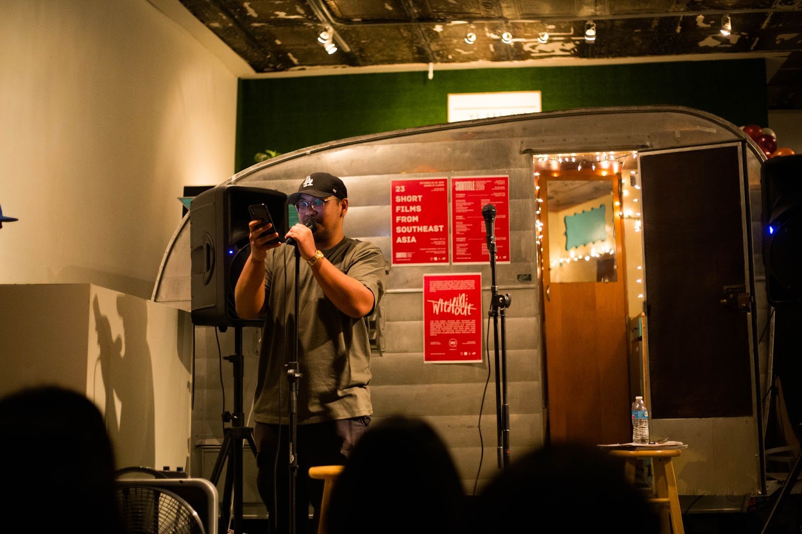 Image: Jofred Estilo performs at Luya at Isa Studios in Pilsen in October 2019. Jofred stands at a microphone reading off of a phone. There is what looks like an air stream-style trailer in the background. Photo by Caroline Olsen.