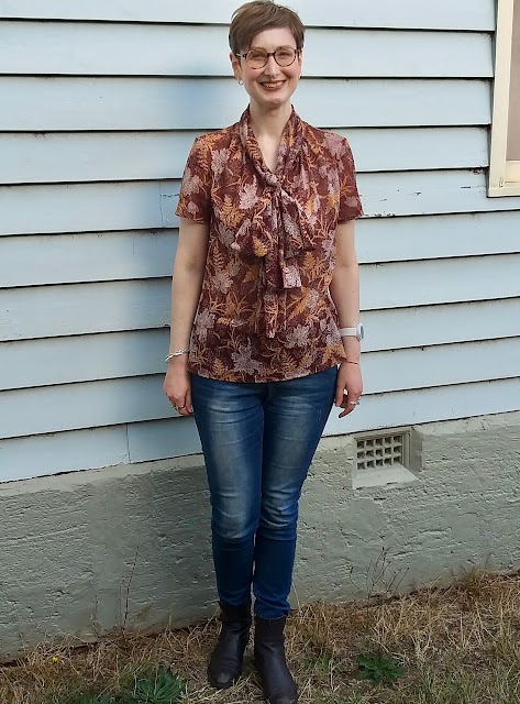 Siobhan stands in front of a blue weatherboard wall. She wears an earthy brown, fern-print short sleeve blouse with comically large bow at neck, blue slim jeans and brown boots. She is smiling.